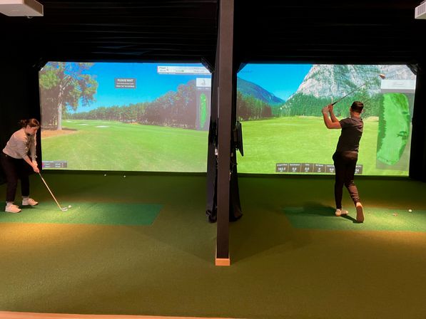 Two golfers in side-by-side indoor golf simulator bays practicing swings on green turf mats with large projected fairway screens showing a virtual course and shot distance stats.