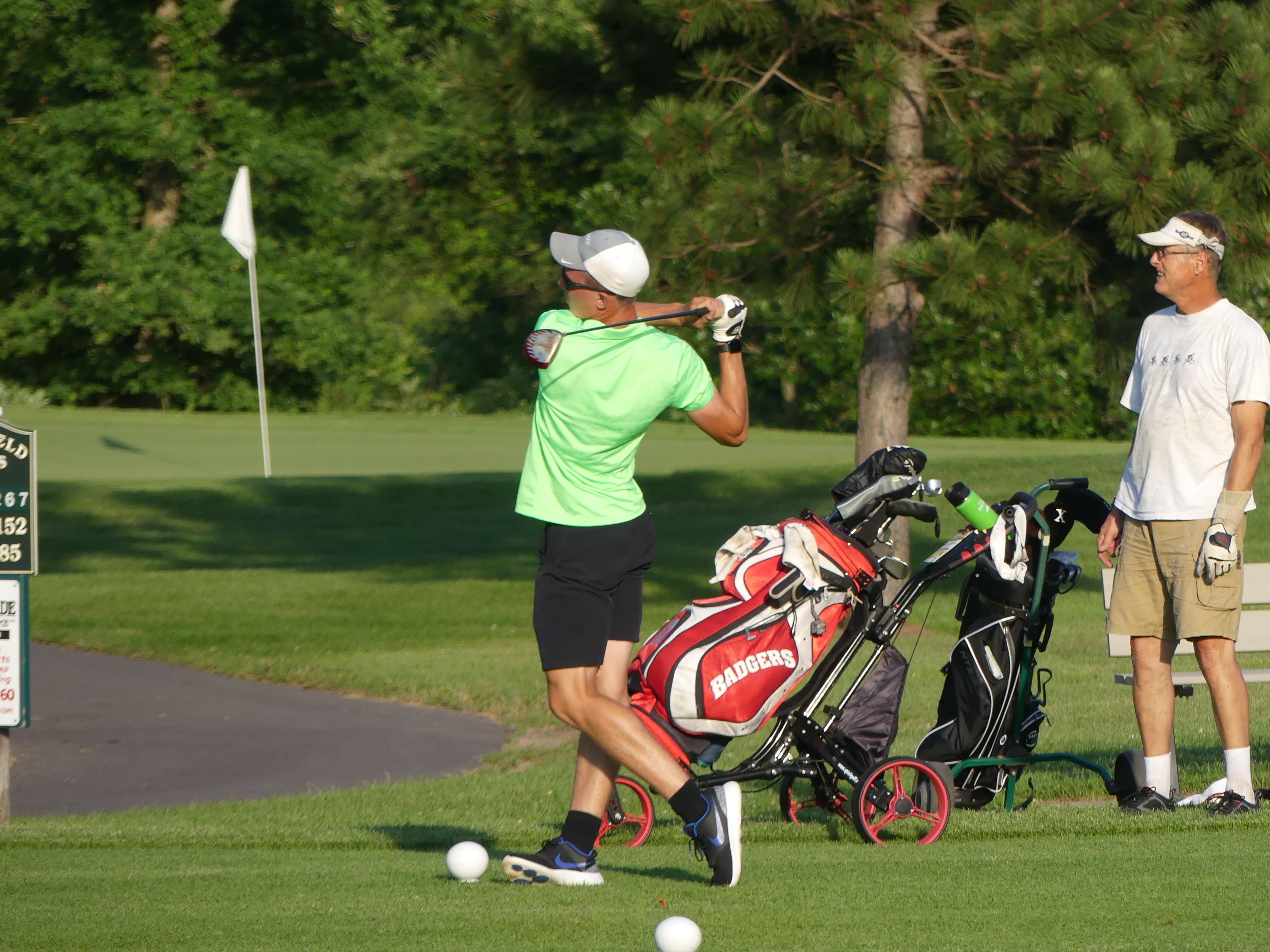 Golfer in neon green shirt mid-swing on a tee box at a tree-lined golf course, red-and-white golf bag on a push cart nearby and a companion watching by the flagstick.