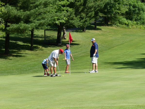 Adult and two children putting on a sunlit golf course putting green by a red flag, surrounded by leafy trees on a summer day