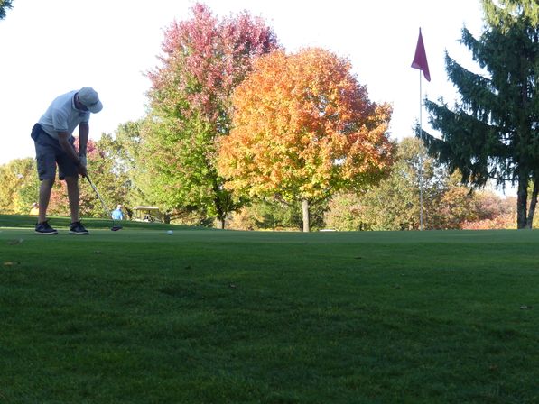 Golfer putting on a sunlit green toward a red flag, framed by vibrant orange and red autumn trees and a nearby golf cart.