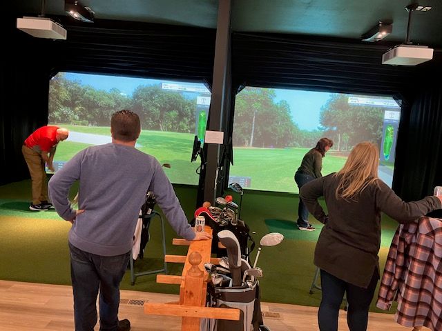 Group practicing at an indoor golf simulator bay: two large screens show a virtual fairway, golfers preparing swings, and a rack of clubs in the foreground.
