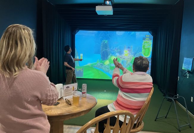 Three adults clap and cheer as a player points at a colorful projected course in an indoor virtual golf simulator bay, with a wooden table and drinks in the foreground.