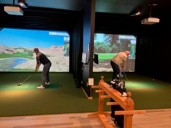 Two golfers practicing in adjacent indoor golf simulator bays with large projected virtual courses on screens, green turf mats, and a wooden club rack with clubs in the foreground.