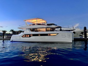 Sleek white luxury catamaran yacht number 53 docked at a marina at twilight, glowing upper deck and cabin lights reflecting golden streaks on calm blue water with palm trees and nearby boats in the background.