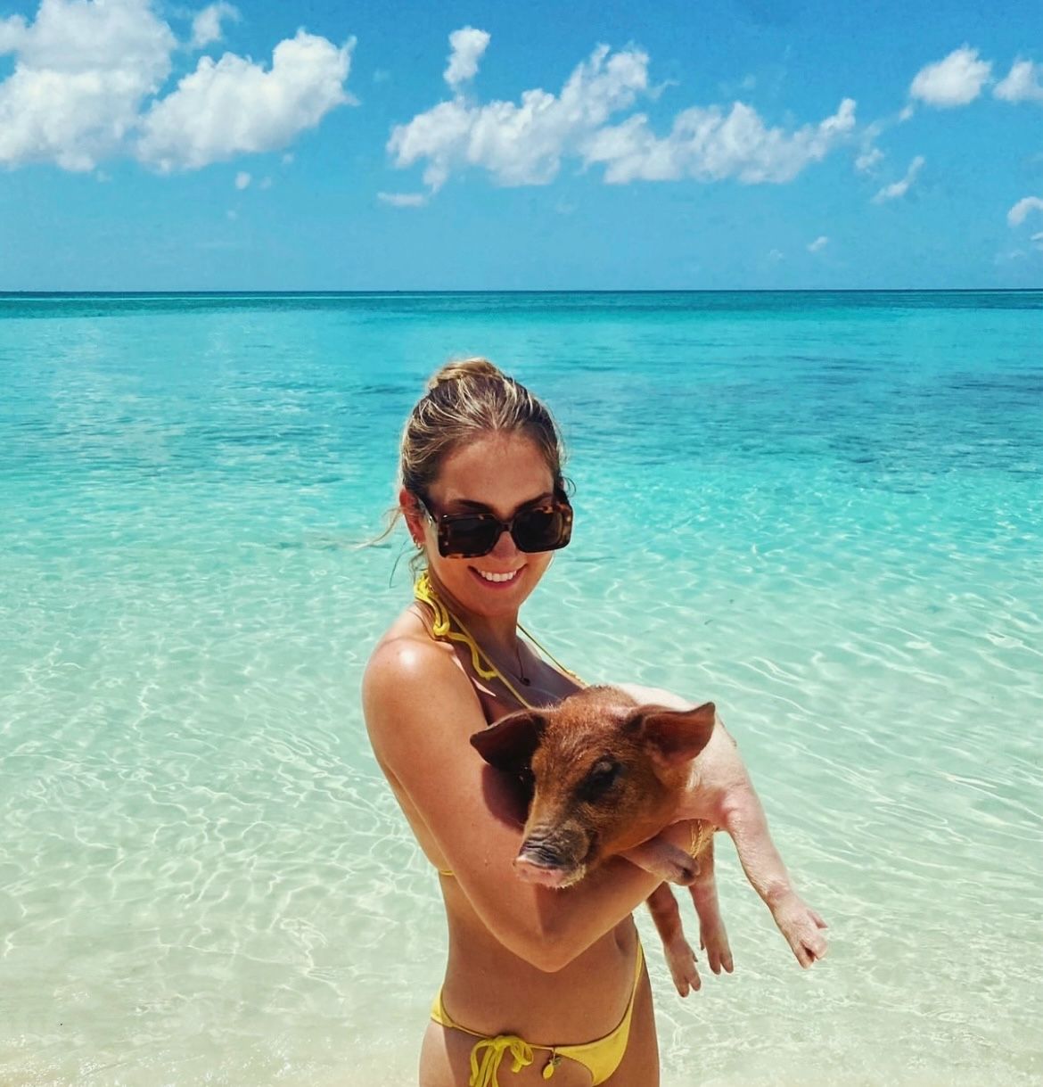 Smiling person in a yellow bikini and sunglasses holding a small piglet in crystal-clear turquoise water at a sunny tropical beach with blue sky and clouds.