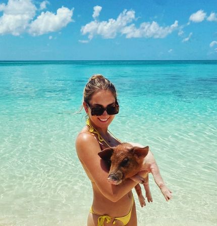 Smiling person in a yellow bikini and sunglasses holding a small piglet in crystal-clear turquoise water at a sunny tropical beach with blue sky and clouds.