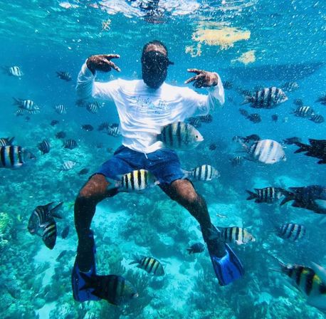 Snorkeler in a white rash guard and blue fins suspended in clear turquoise water, surrounded by striped tropical fish over a vibrant coral reef — tropical coral reef snorkeling scene.