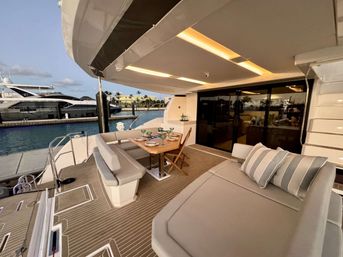 Luxury yacht aft deck at a marina with teak-style decking, L-shaped cushioned dining bench and table set for four, folding chairs, padded sunbed with striped pillows, sliding glass doors to the interior, and nearby moored yachts in evening light.