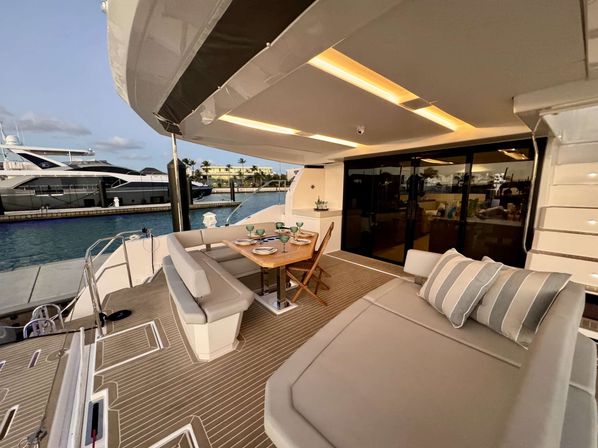 Luxury yacht aft deck at a marina with teak-style decking, L-shaped cushioned dining bench and table set for four, folding chairs, padded sunbed with striped pillows, sliding glass doors to the interior, and nearby moored yachts in evening light.