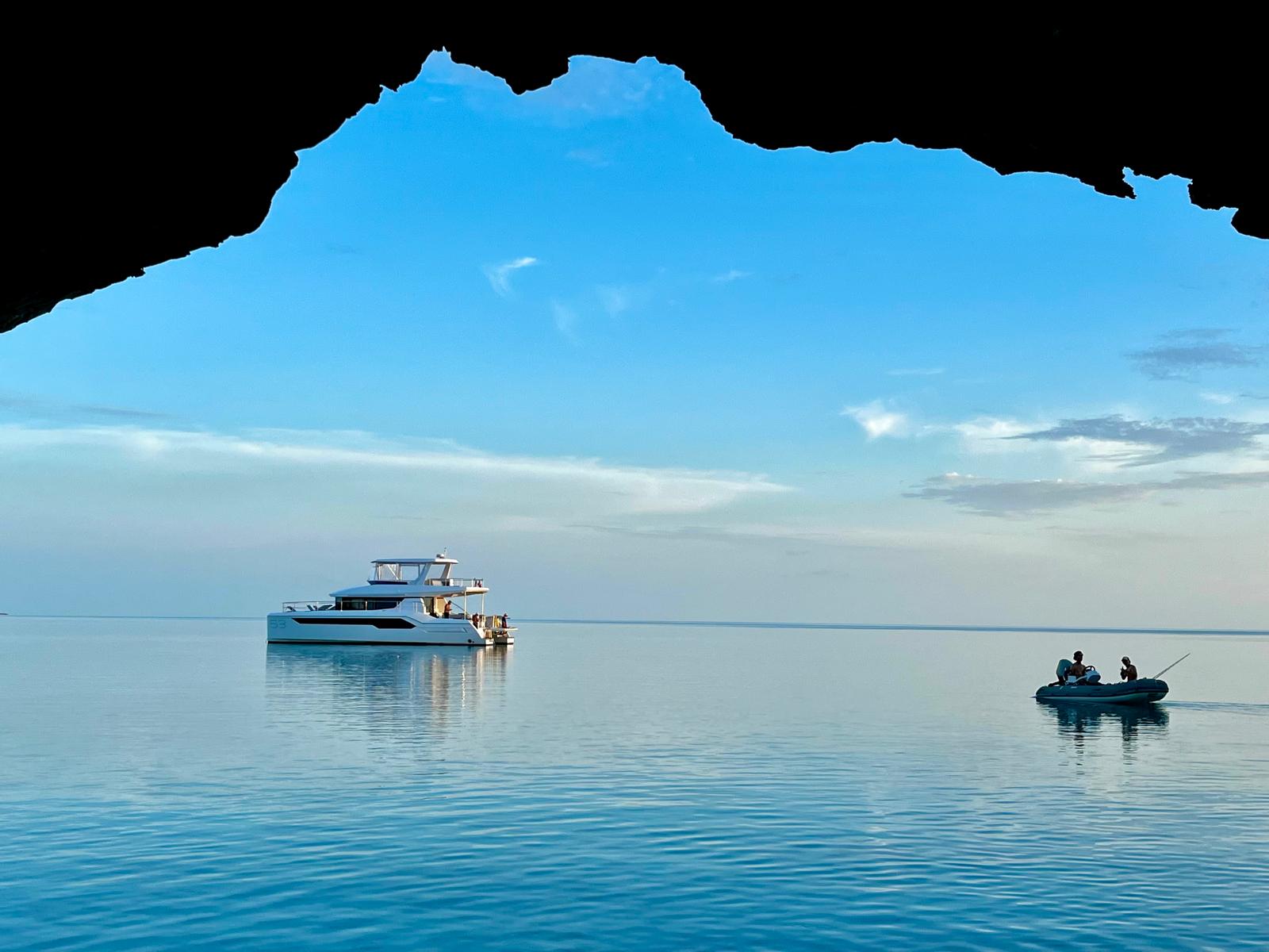 Sea cave silhouette framing a calm blue ocean with a white yacht anchored left and a small inflatable dinghy with two people fishing on the right under a clear sky — postcard‑calm coastal scene.