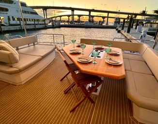 Sunset yacht dining setup at a marina: wooden table with plates and green glass goblets, cushioned bench seating and folding chairs overlooking water and a bridge.