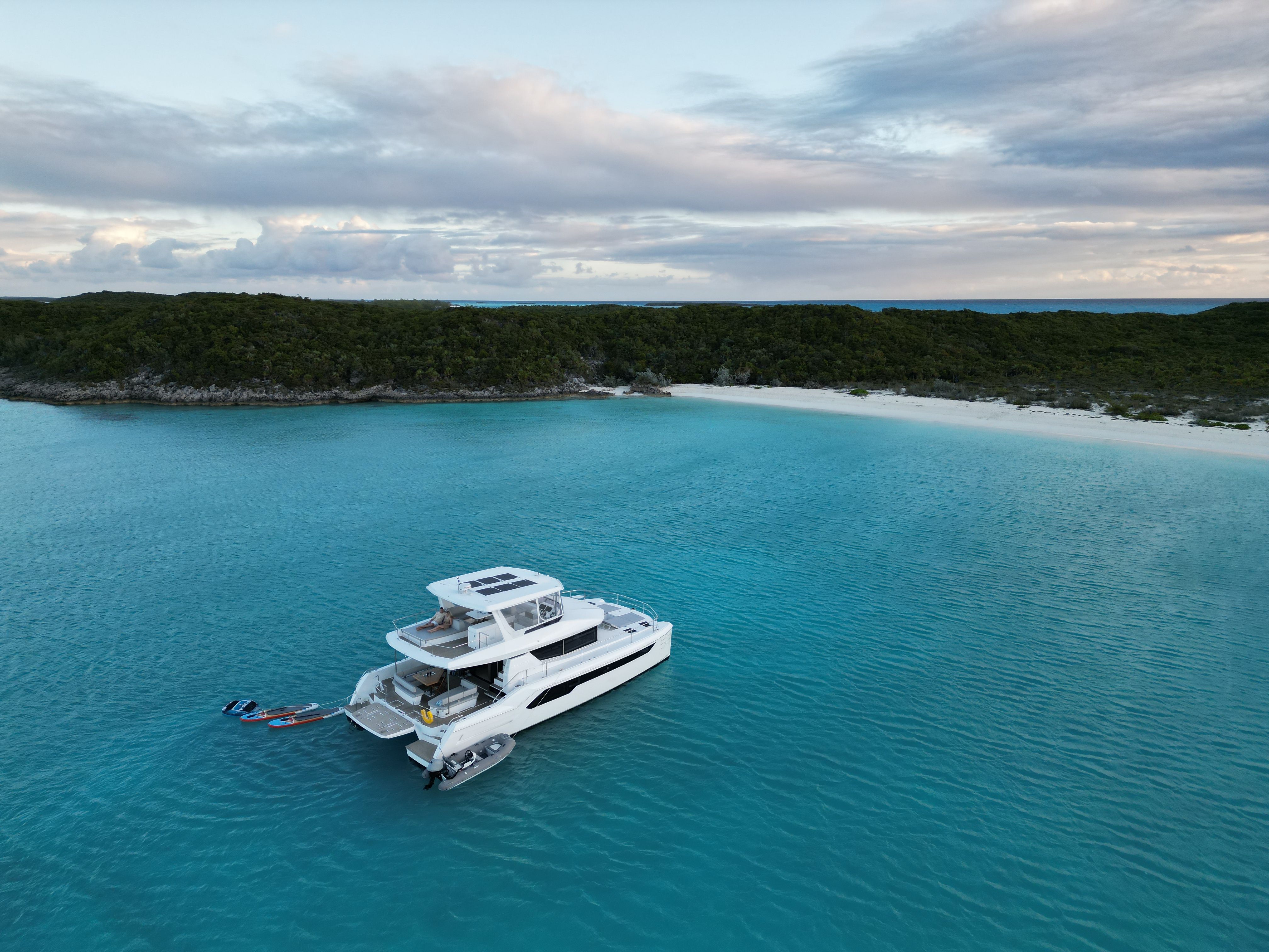 Aerial view of a white luxury yacht anchored in turquoise tropical waters off a white-sand beach and green island, with paddleboards and a small tender trailing at the stern.