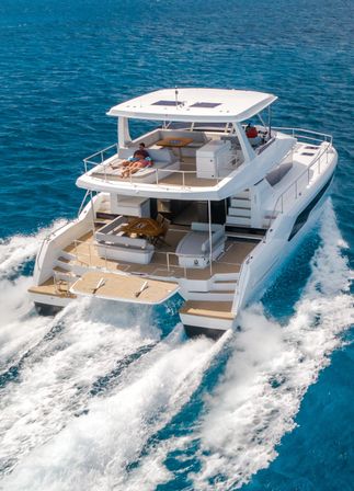 Luxury white catamaran cruising on deep blue ocean, twin-hull wake and frothy waves, rear deck lounge with sunbathers and shaded upper helm