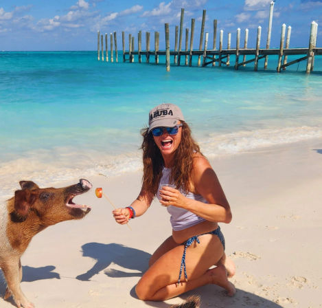 Laughing woman kneeling on a white-sand tropical beach offering a fruit skewer to a pig, with turquoise water and a wooden pier in the background.
