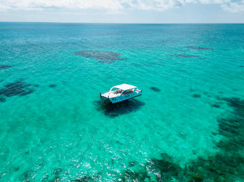 Aerial view of a white catamaran floating over clear turquoise tropical water with dark coral patches visible below and a calm horizon under a partly cloudy sky.