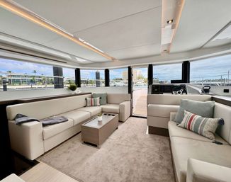 Sunlit modern luxury yacht salon with beige L-shaped sofas, coffee table and patterned pillows, panoramic windows offering marina and waterfront views