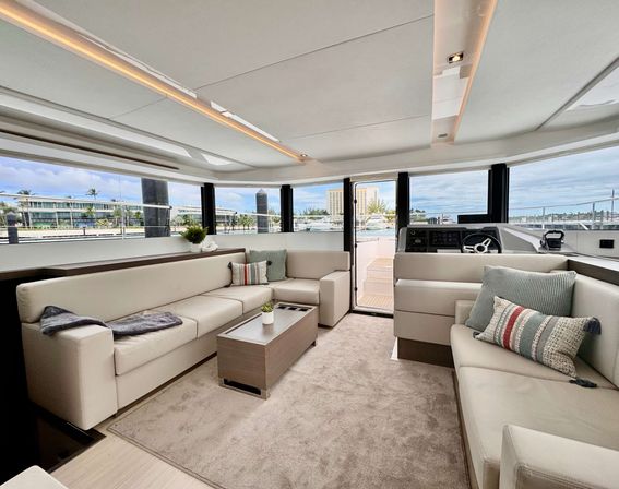 Sunlit modern luxury yacht salon with beige L-shaped sofas, coffee table and patterned pillows, panoramic windows offering marina and waterfront views