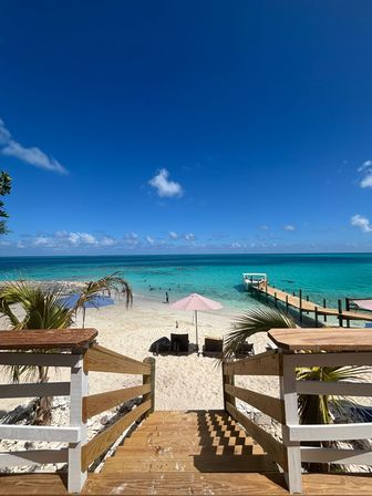 Inviting wooden steps down to a tropical white-sand beach with a pink umbrella, lounge chairs, turquoise water and a pier under a clear blue sky.