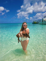 Woman in white swimsuit wearing heart-shaped sunglasses holding a piglet in shallow turquoise tropical water near boats and a dock under a sunny blue sky