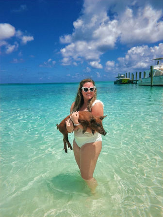 Woman in white swimsuit wearing heart-shaped sunglasses holding a piglet in shallow turquoise tropical water near boats and a dock under a sunny blue sky