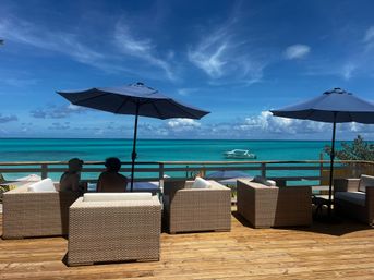 Wooden beachfront deck with wicker lounge chairs and navy umbrellas, two silhouetted people seated, turquoise tropical sea and a small boat beneath a bright blue sky