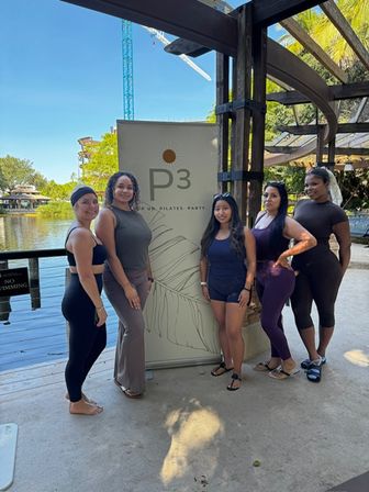 Five women in activewear pose by a lakeside pergola next to a fitness banner — sunny outdoor pilates/fitness meetup with palm trees and a crane in the background.