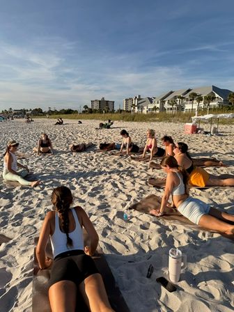 Sunlit beach yoga class: a group stretching on mats across white sand near beachfront condos under a clear blue sky