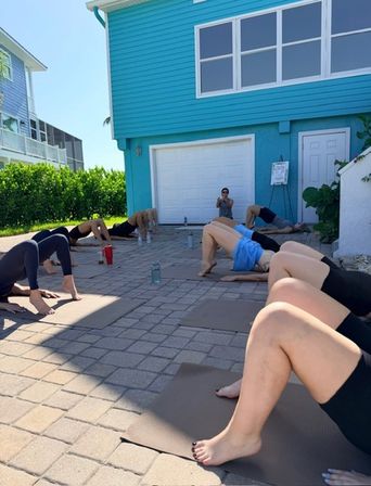 Group yoga class outdoors on exercise mats on a paved driveway in front of a bright turquoise coastal house, participants doing glute-bridge core exercises with water bottles in the sun.