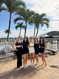 Five women in coordinated black-and-white summer outfits pose on a sunny waterfront patio at a palm-lined marina with boats docked and a bright blue sky.