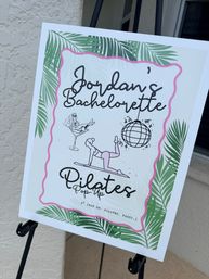 Bachelorette pilates pop-up event sign on an easel with tropical palm-leaf border, pink scalloped frame and playful pilates illustrations and disco ball, displayed outside by a doorway