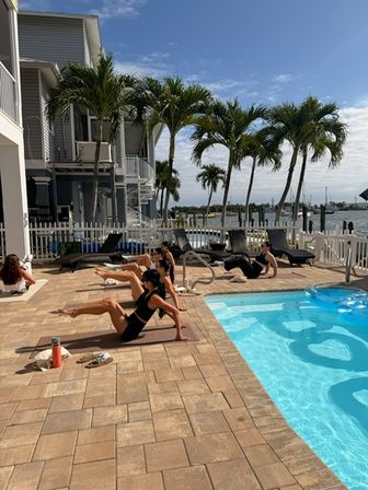 Energetic poolside workout on a sunny tropical waterfront deck — group exercising on mats by a blue pool with palm trees, loungers and marina views.