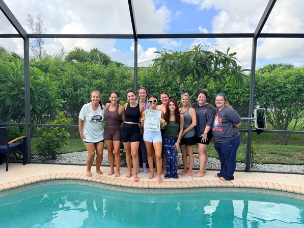 Group of women smiling and posing poolside inside a screened backyard pool enclosure with tropical greenery, one woman holding a bachelorette party sign.