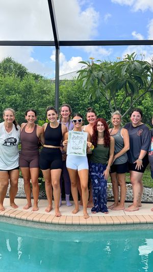 Group of women smiling and posing poolside inside a screened backyard pool enclosure with tropical greenery, one woman holding a bachelorette party sign.