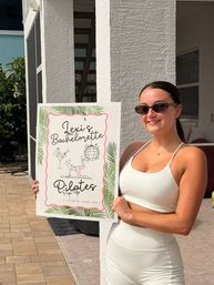 Person in a white workout set and sunglasses smiling on a sunny backyard patio, holding a decorative sign for a bachelorette Pilates pop-up event.