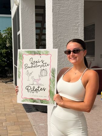 Person in a white workout set and sunglasses smiling on a sunny backyard patio, holding a decorative sign for a bachelorette Pilates pop-up event.