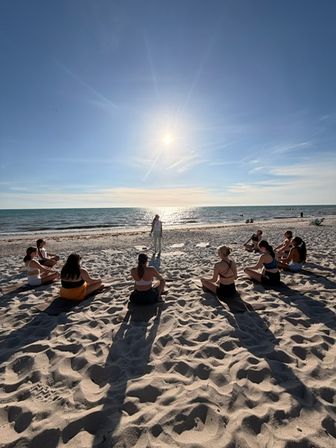 Oceanfront beach yoga—a circle of people seated on white sand facing an instructor by the sparkling sea under a bright late-afternoon sun.