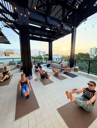 Rooftop sunset yoga class on a covered terrace — group practicing boat pose on mats beneath string lights with palm trees and a coastal city skyline in the background.