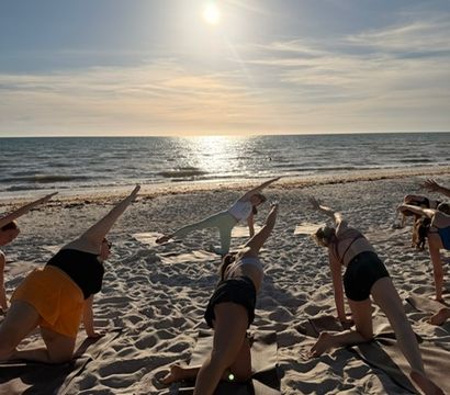 Group practicing beach yoga at sunset on a sandy shore, participants in side-plank poses facing the sun reflecting on the ocean.