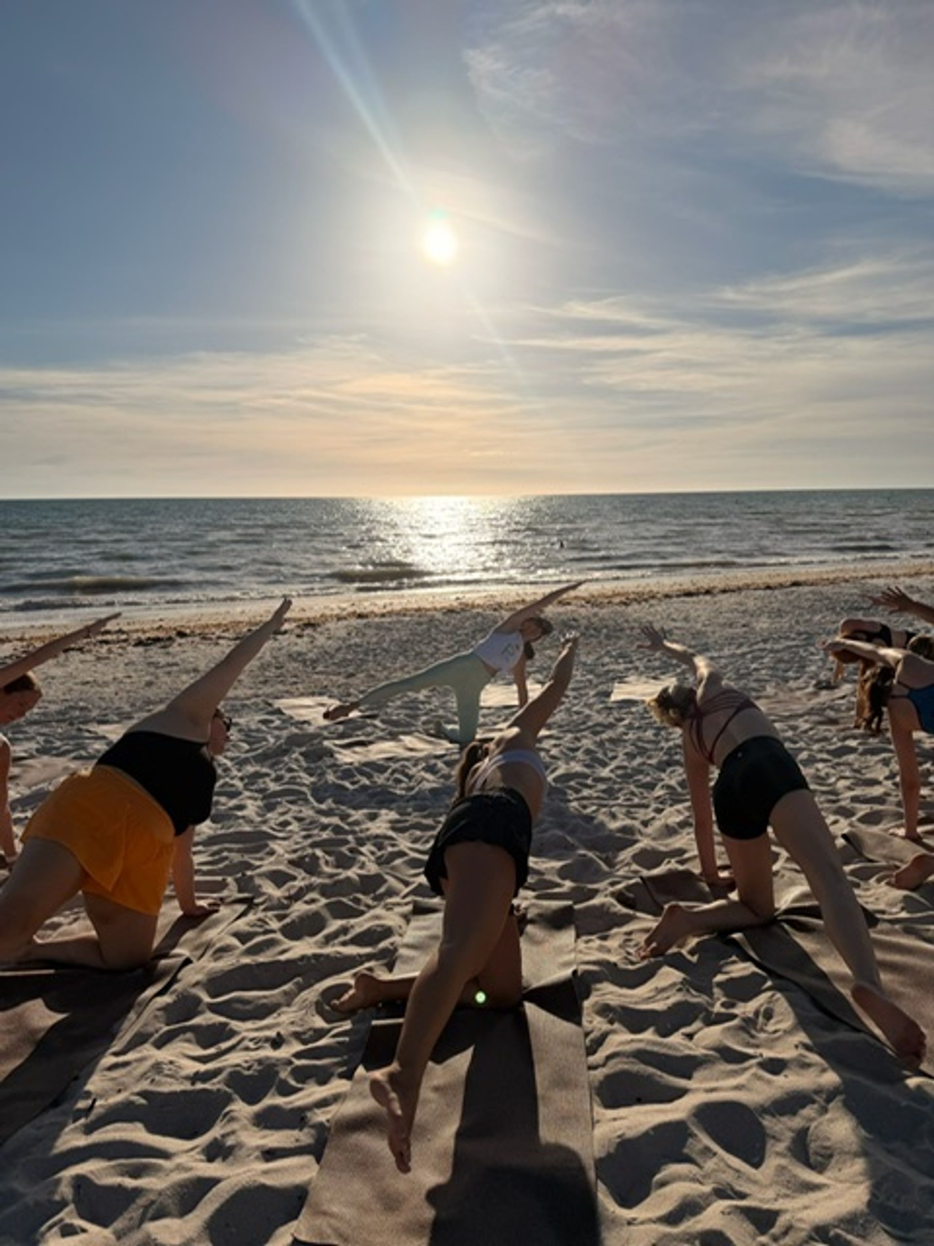Group practicing beach yoga at sunset on a sandy shore, participants in side-plank poses facing the sun reflecting on the ocean.