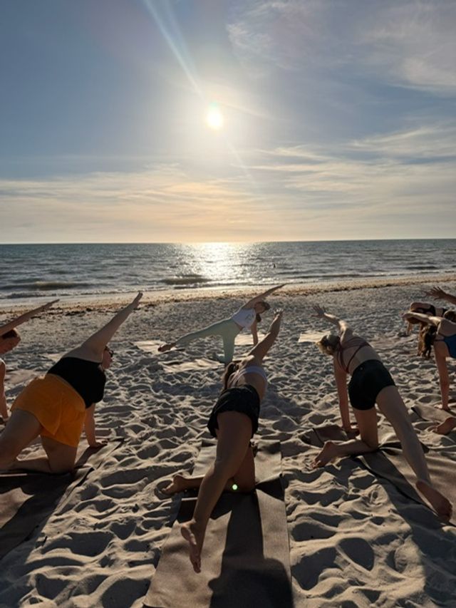 Group practicing beach yoga at sunset on a sandy shore, participants in side-plank poses facing the sun reflecting on the ocean.