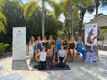 Group of women in colorful activewear posing on a sunny tropical poolside patio beneath palm trees at an outdoor fitness/yoga event.