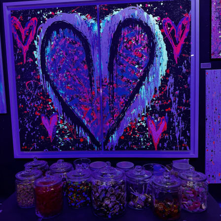 UV blacklight neon heart painting on split panels — dripping turquoise, pink and purple splatter with small hearts — row of glass candy jars on a table below