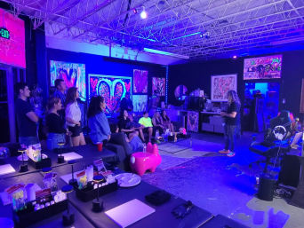 Neon-lit warehouse art studio with vivid heart and abstract paintings, a seated group watching an artist give a live painting demo in a relaxed gallery-style workshop.