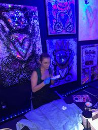 Artist in UV-lit art studio decorating a denim jacket under blacklight, surrounded by neon fluorescent paintings of hearts and bears.