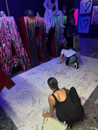 Interactive indoor art studio scene: two people crouched signing a large white floor canvas in front of oversized black letters spelling "ART" splattered with neon paint under UV lighting, colorful graffiti panels and mannequins in the background.