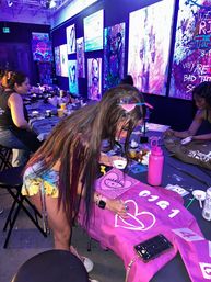Neon-lit art studio DIY paint night with people customizing pink denim jackets; woman painting a white heart on a jacket amid colorful abstract canvases on the walls.