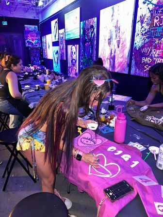 Neon-lit art studio DIY paint night with people customizing pink denim jackets; woman painting a white heart on a jacket amid colorful abstract canvases on the walls.