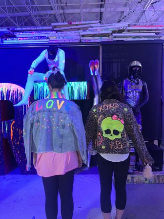 Two people seen from behind wearing custom neon-painted jackets — a denim jacket reading 'LOVE' and a leopard-print jacket with a green skull — standing in a UV-lit warehouse art installation with fluorescent-painted mannequins and dripping neon backdrops.