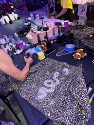 Leopard-print denim jacket on a craft workshop table being customized with a sparkly glitter snake and stars, surrounded by paints, brushes, tape and small storage baskets in an indoor DIY studio.