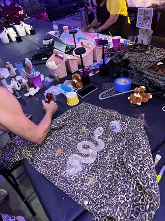 Leopard-print denim jacket on a craft workshop table being customized with a sparkly glitter snake and stars, surrounded by paints, brushes, tape and small storage baskets in an indoor DIY studio.